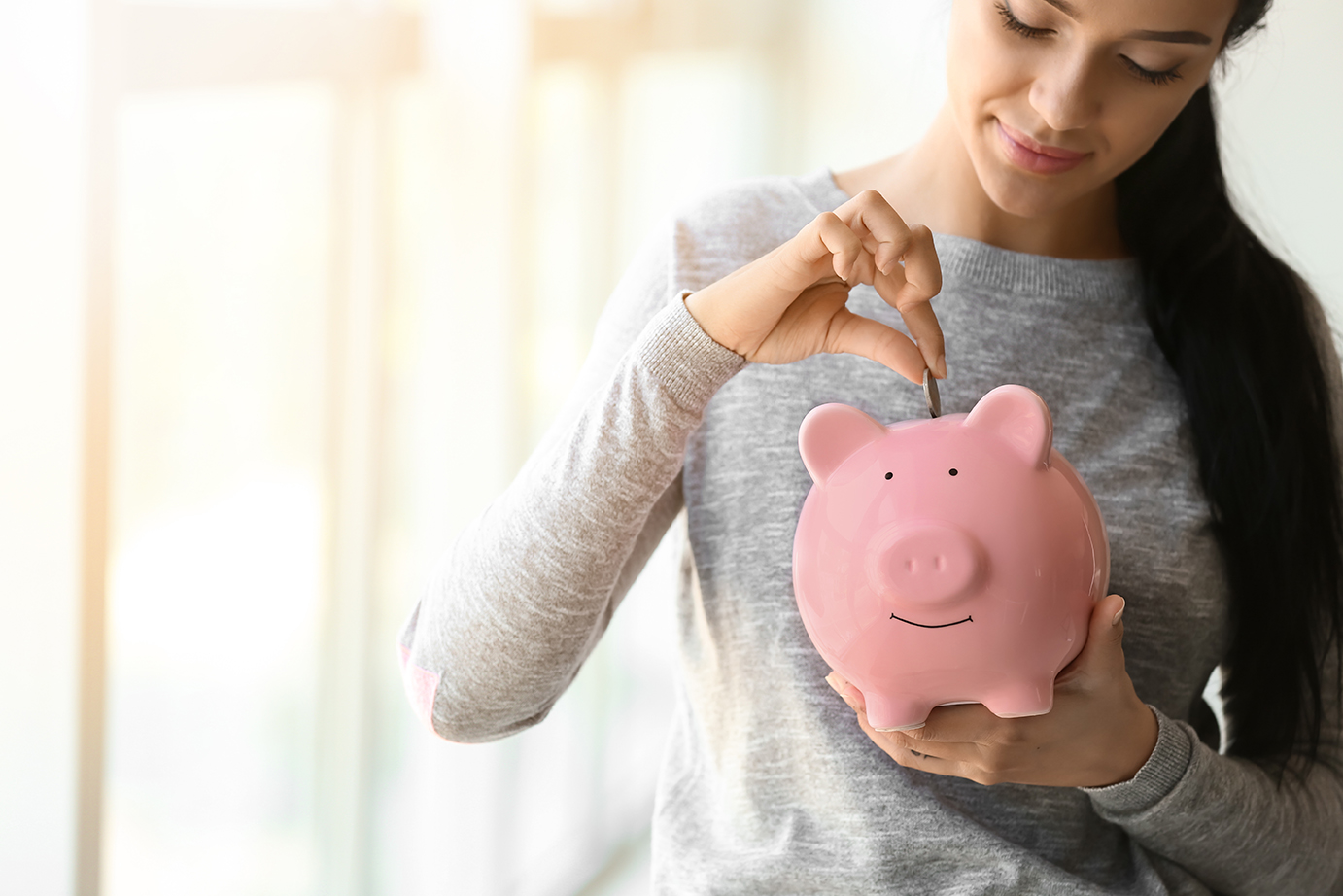 Young,Woman,Putting,Money,Into,Piggy,Bank,Near,Window